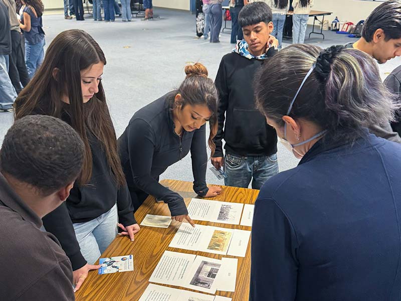 four high school students engaging at a table station pointing to a document with a photo